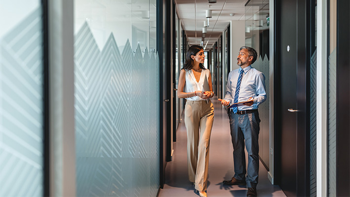 Two individuals standing in an office hallway, engaged in conversation near a set of closed doors.
