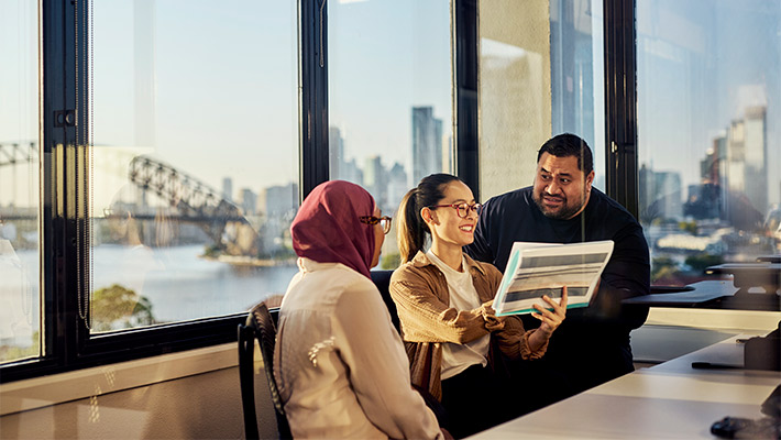 Three people sit in an office with large windows overlooking a harbor and city skyline, collaborating while looking at a document.