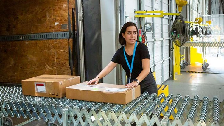 A young woman employed at a fulfillment center using an extendable conveyor belt to load boxes and packages.
